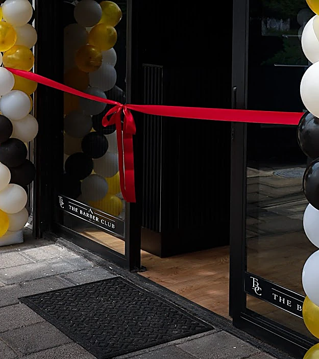 Puerta de vidrio de la barbería con un listón rojo de inauguración y globos a los lados