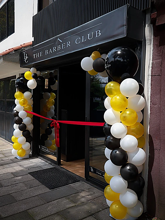 Fachada de The Barber Club el día de la inauguración: listones negros, toldo con el nombre, listón rojo en la puerta y columnas de globos negros, dorados y blancos