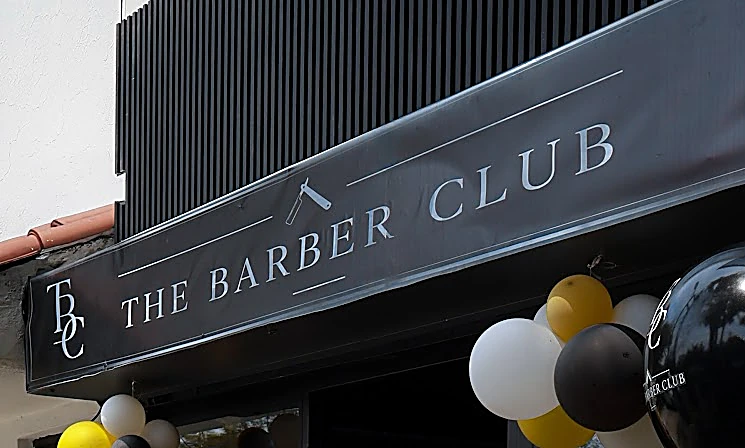 Toldo negro con el monograma TBC y el nombre The Barber Club en letras plateadas, bajo los listones de la fachada